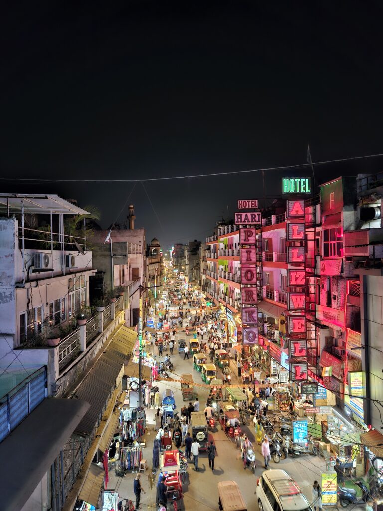 Drukke straat in New Delhi bij nacht met neonreclames, hotels, riksja’s, marktkraampjes en een bruisende menigte.