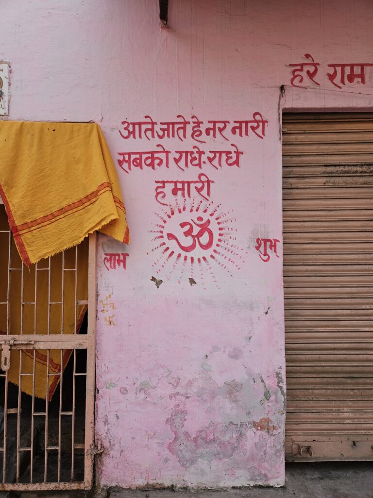 Roze muur in Agra met Hindi-inscripties, een groot Om-symbool en een geel doek naast een metalen hek.