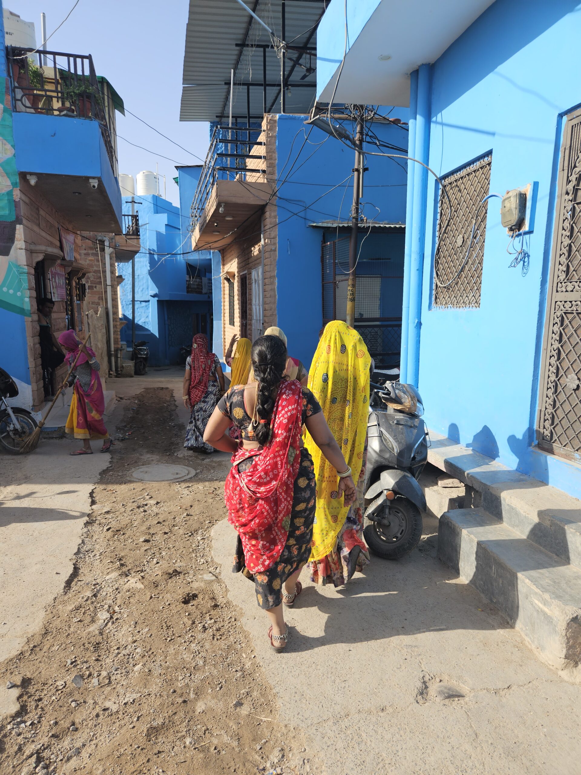 Vrouwen in kleurrijke sari’s lopen door een smalle straat met felblauwe huizen in India.