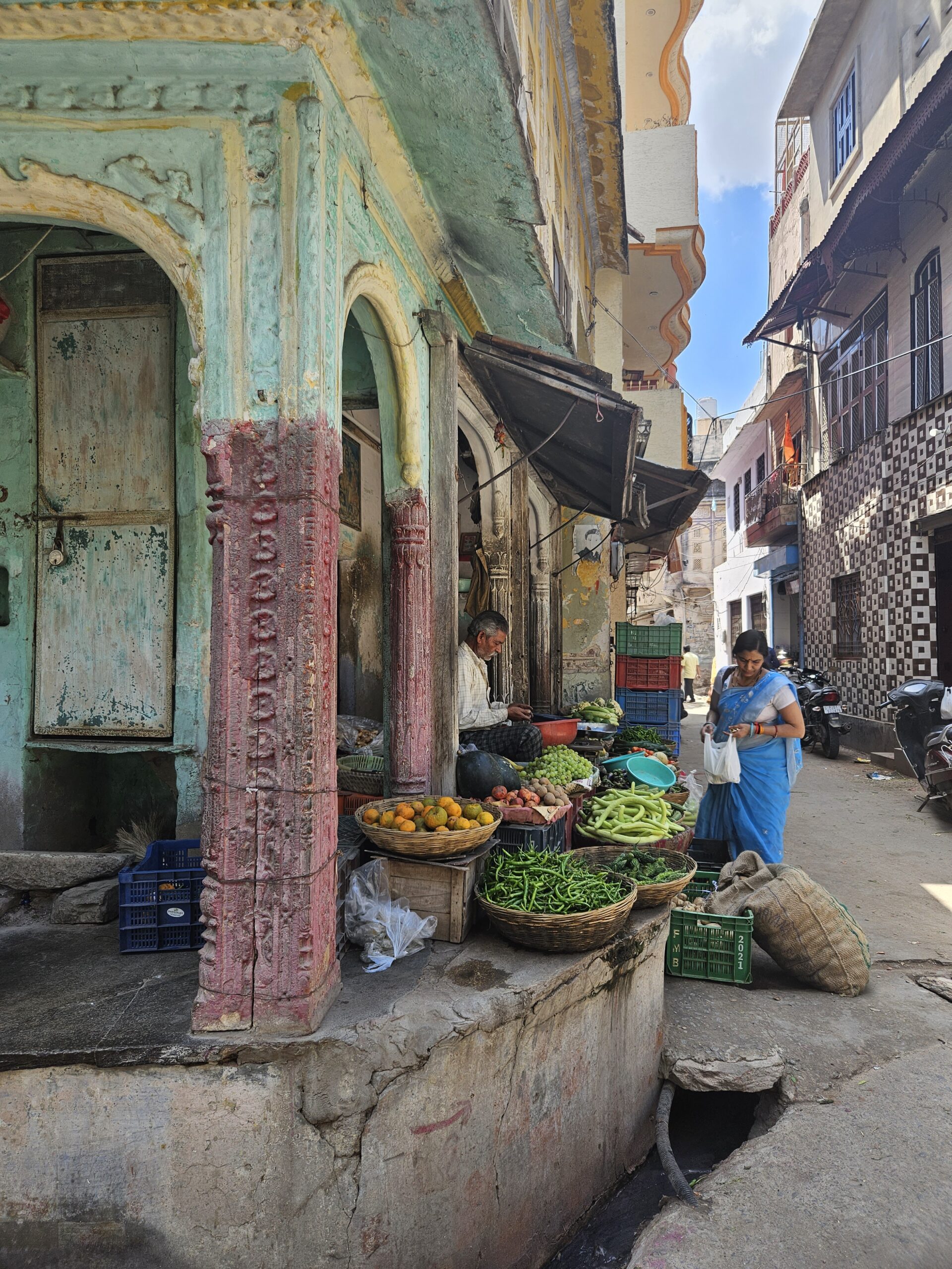 Groentekraam in een smalle straat in India met kleurrijke groenten en een vrouw in een blauwe sari die inkopen doet.