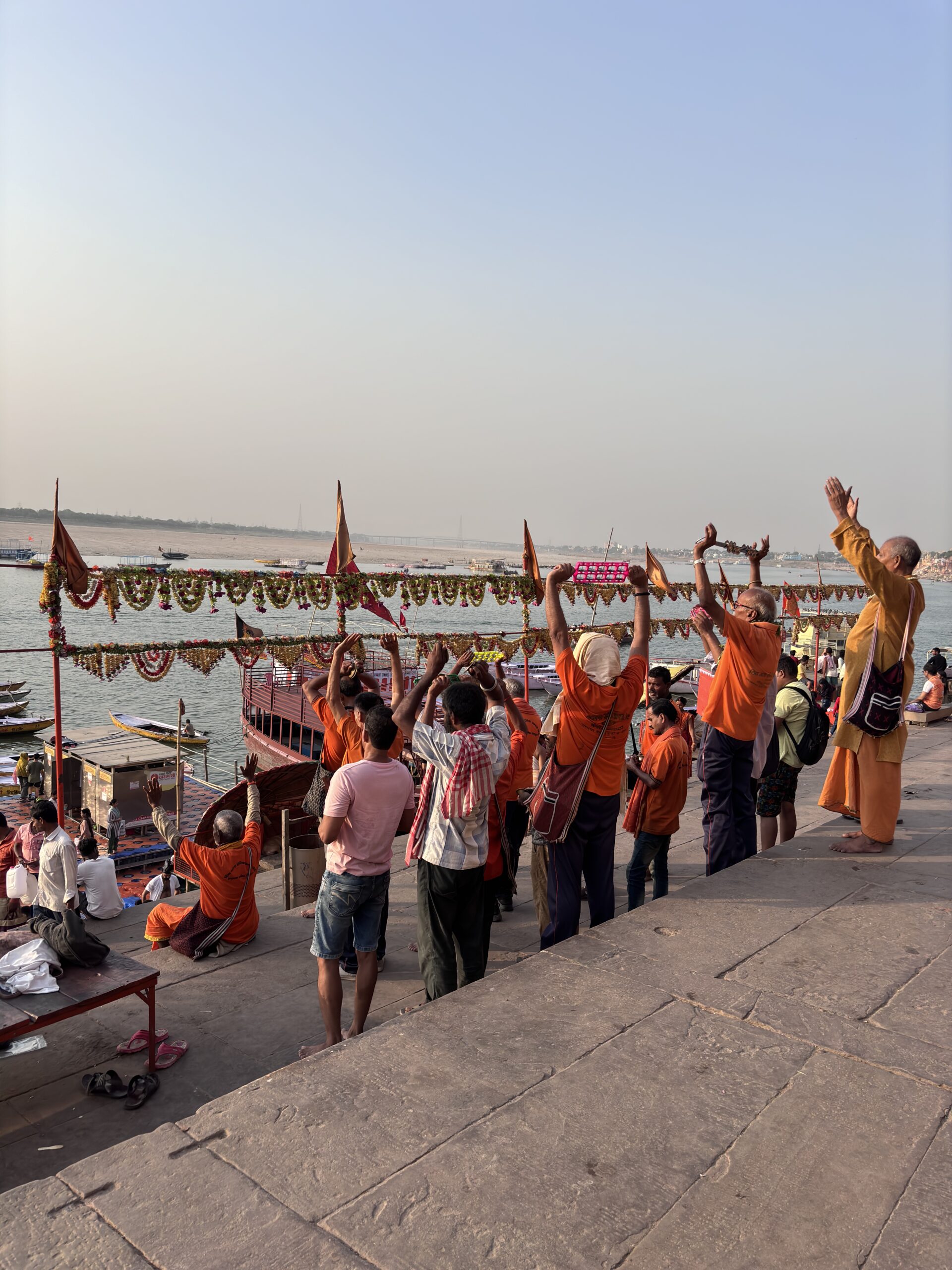 Groep gelovigen in oranje kleding bidt en heft armen omhoog tijdens ritueel aan de Ganges in Varanasi, India.