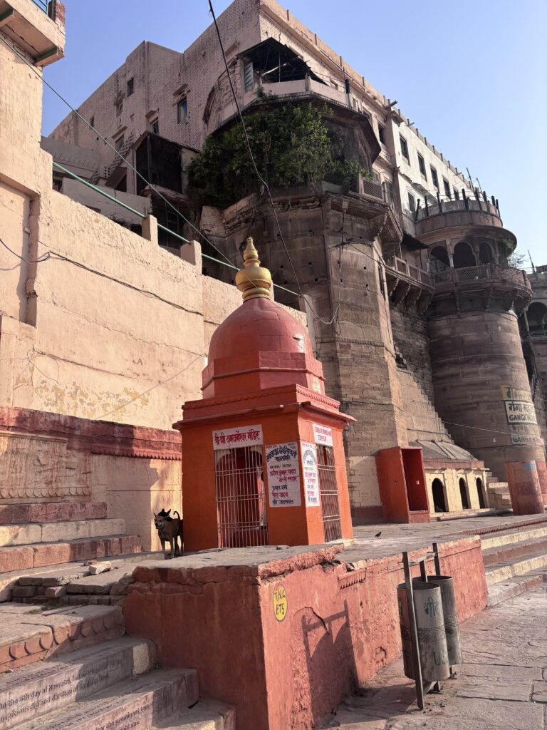 Kleine rode tempel met gouden koepel langs de ghats van Varanasi, India, met historische gebouwen op de achtergrond.