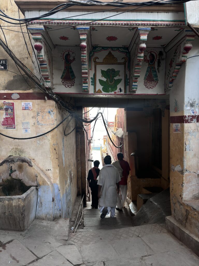 Mensen lopen door een smalle doorgang met schilderingen en zicht op de ghats in Varanasi, India.