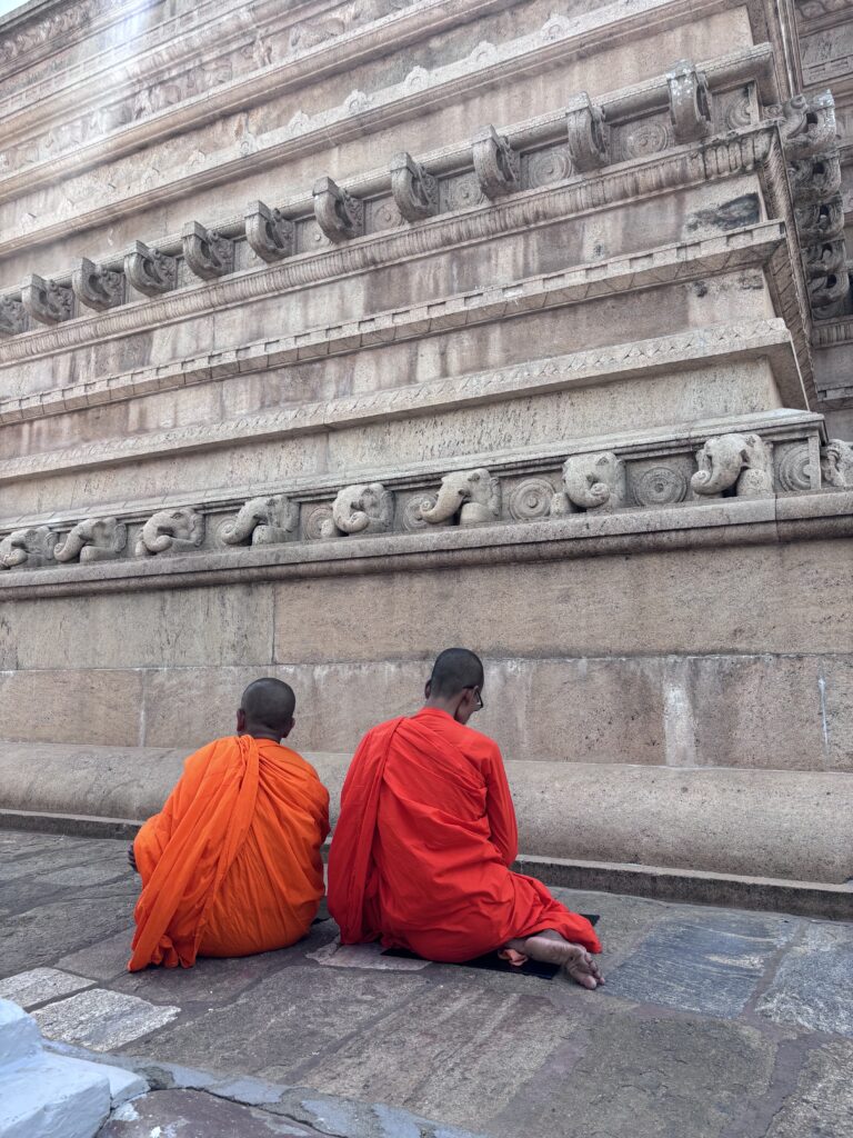 Twee boeddhistische monniken in oranje en rode gewaden bidden bij een oude stenen tempelmuur.