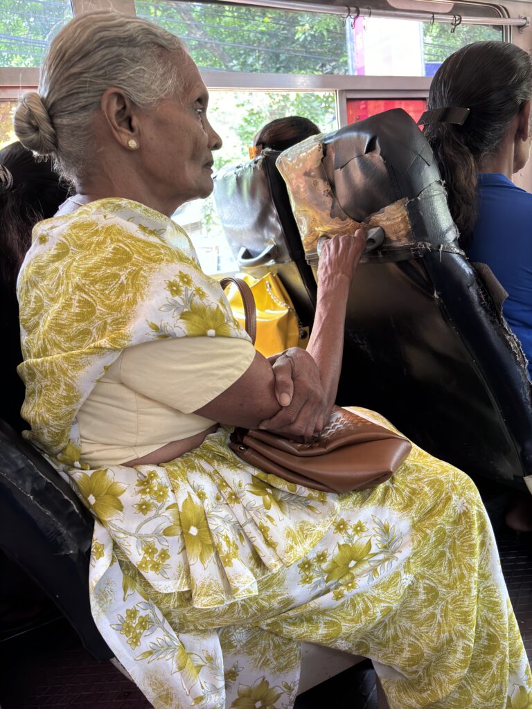 Oudere vrouw in een wit-gele sari zit in een bus, met een tas op schoot en hand op de stoel voor haar.