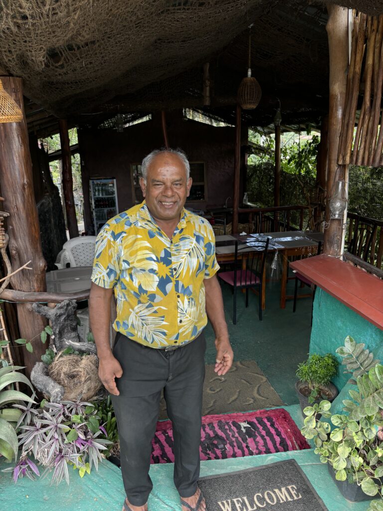 Man in kleurrijk geel-blauw overhemd staat lachend bij de ingang van een restaurant met planten en decoratie.