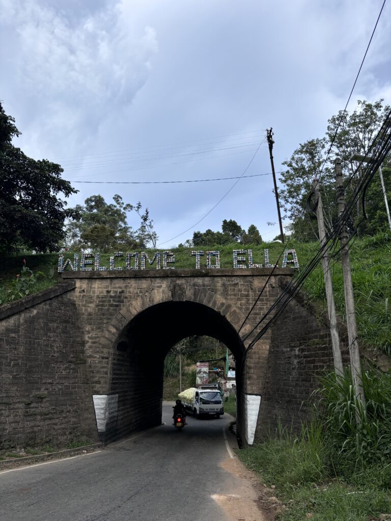 Boogbrug met opschrift "Welcome to Ella" boven een doorgang waar een motor en busje passeren.