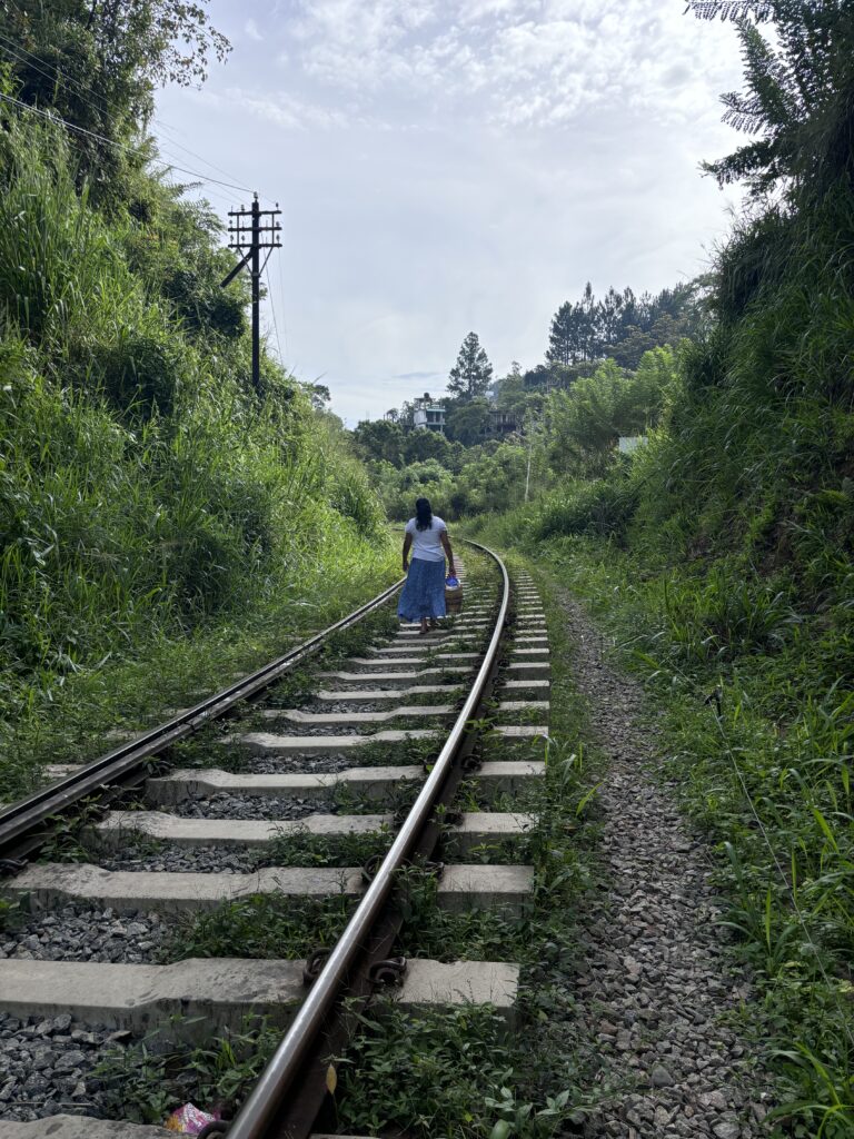 Vrouw in witte blouse en blauwe rok loopt met mand over een spoorlijn door groen landschap in Ella.