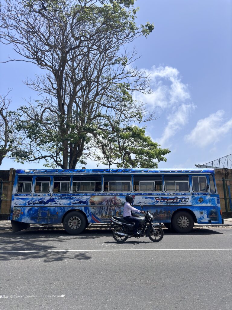 Blauwe bus met kleurrijke muurschildering staat geparkeerd langs de weg, terwijl een man op een motor passeert.