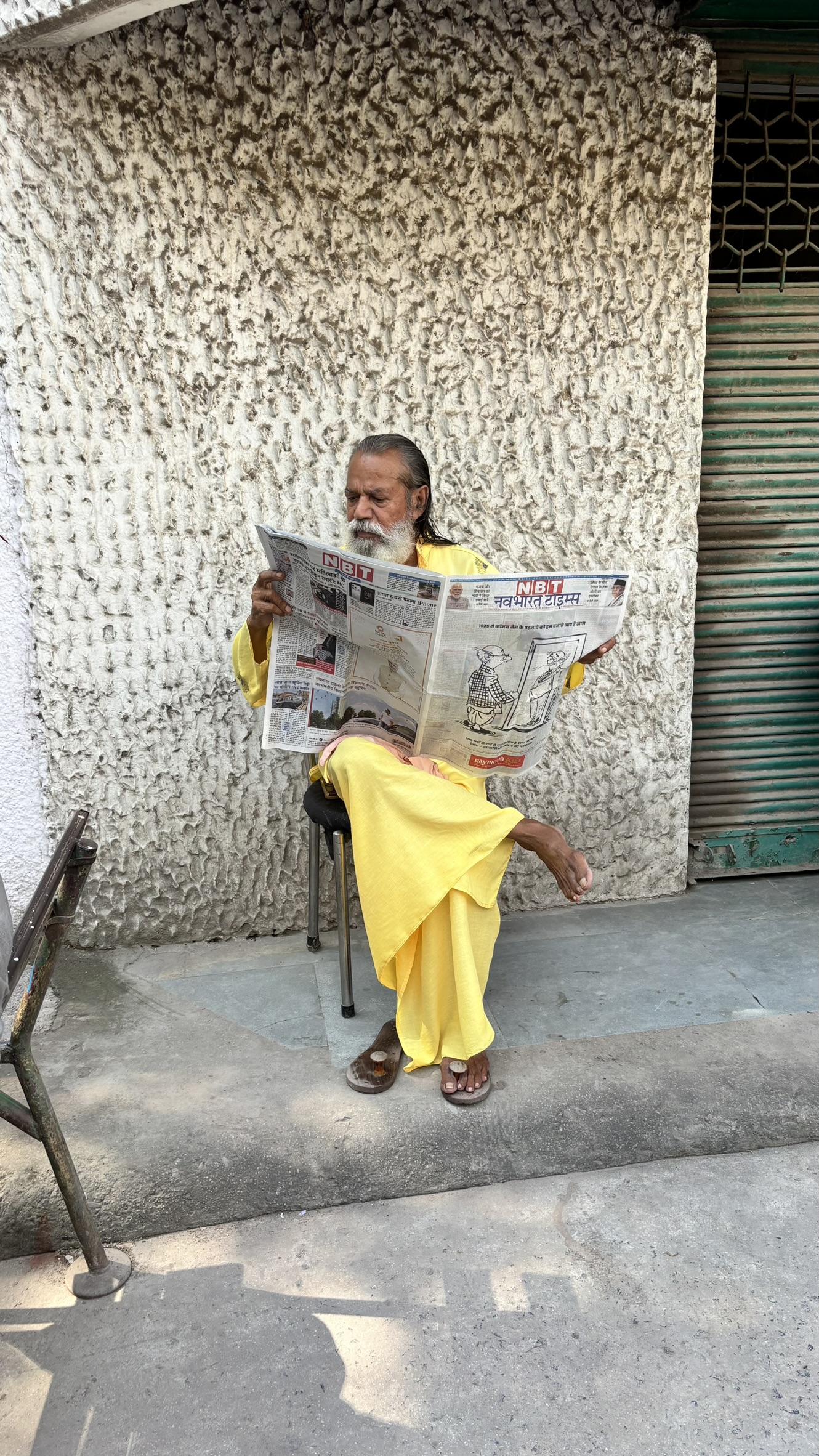 Oudere man in gele gewaden leest een Indiase krant terwijl hij op een krukje zit langs een straatmuur in India.